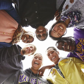 Seven students forming a group circle outside on campus while looking down with smiles as they pose for a photo 