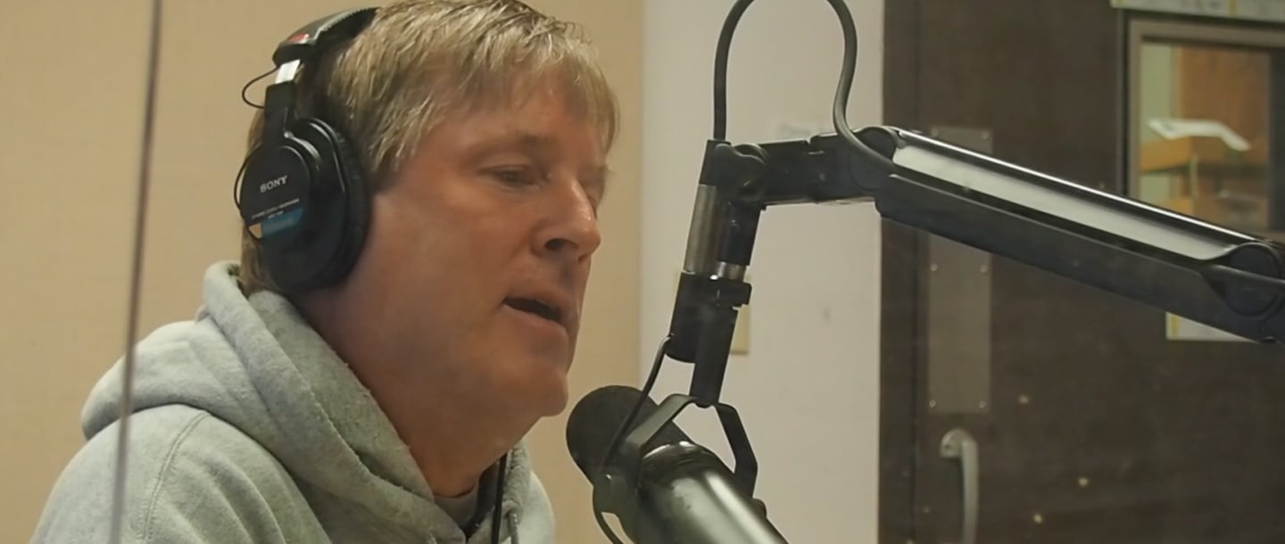 Steve Fligge with a hoodie on, the Supreme Ruler of Sound, speaking into the microphone inside of the KMSU Radio station studio