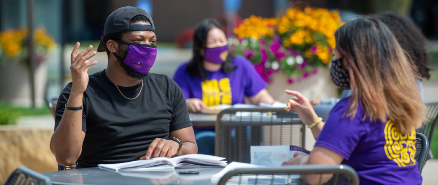 students sitting down at tables outside on campus talking and studying wearing masks