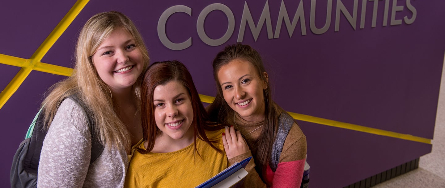Three students standing close to each other for a picture in front of a purple Learning Communications wall