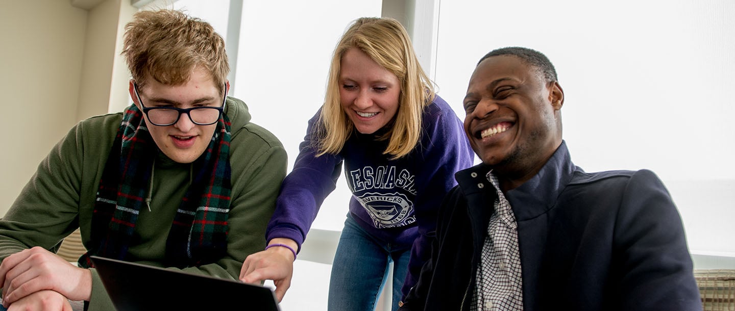 A student pointing at the screen of a computer for two other students while one student on the right is laughing