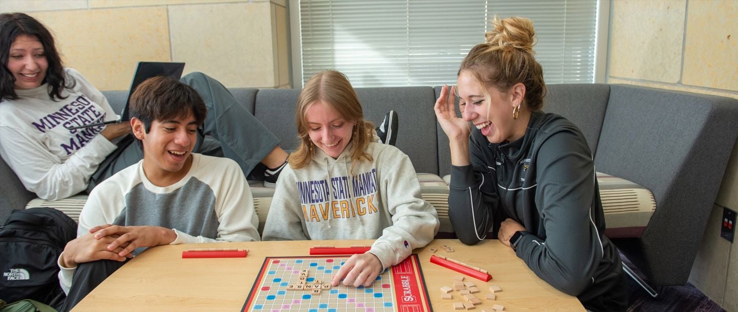 Four students playing scrabble together on a table in the residence hall lobby
