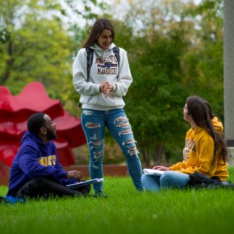 Three students, one standing and two sitting, on the campus lawn talking and studying