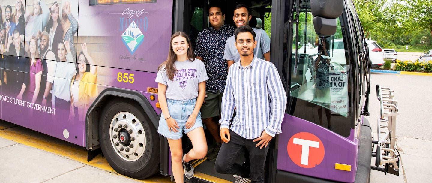 Four students standing in front of a Minnesota State Mankato University bus and posing at the entrance
