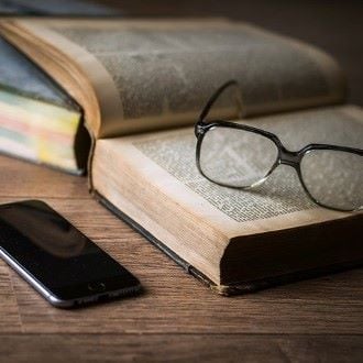 An open book with reading glasses on top next to a cell phone on a table