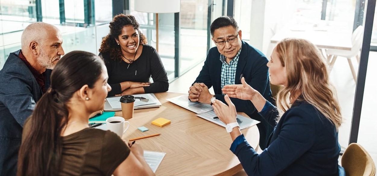 2 women and 2 men are sitting down around a table and listening to another woman speak on the other of the table