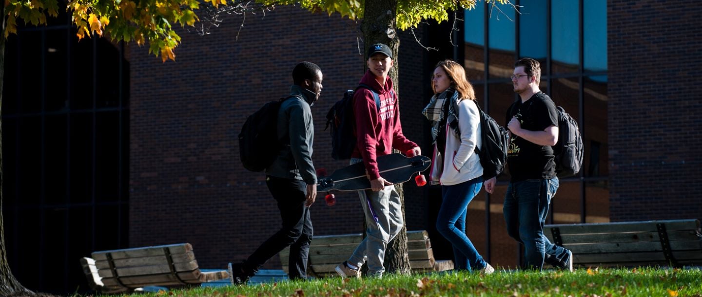 Four students walking outside on campus with backpacks and one student is holding a skateboard