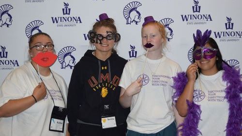 A group of the College Tour Mania students posing with silly props in front of a wall that says Winona State University