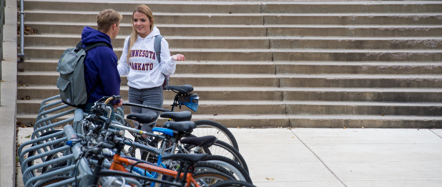 Two students talking outside on campus at a bicycle rack near some stairs