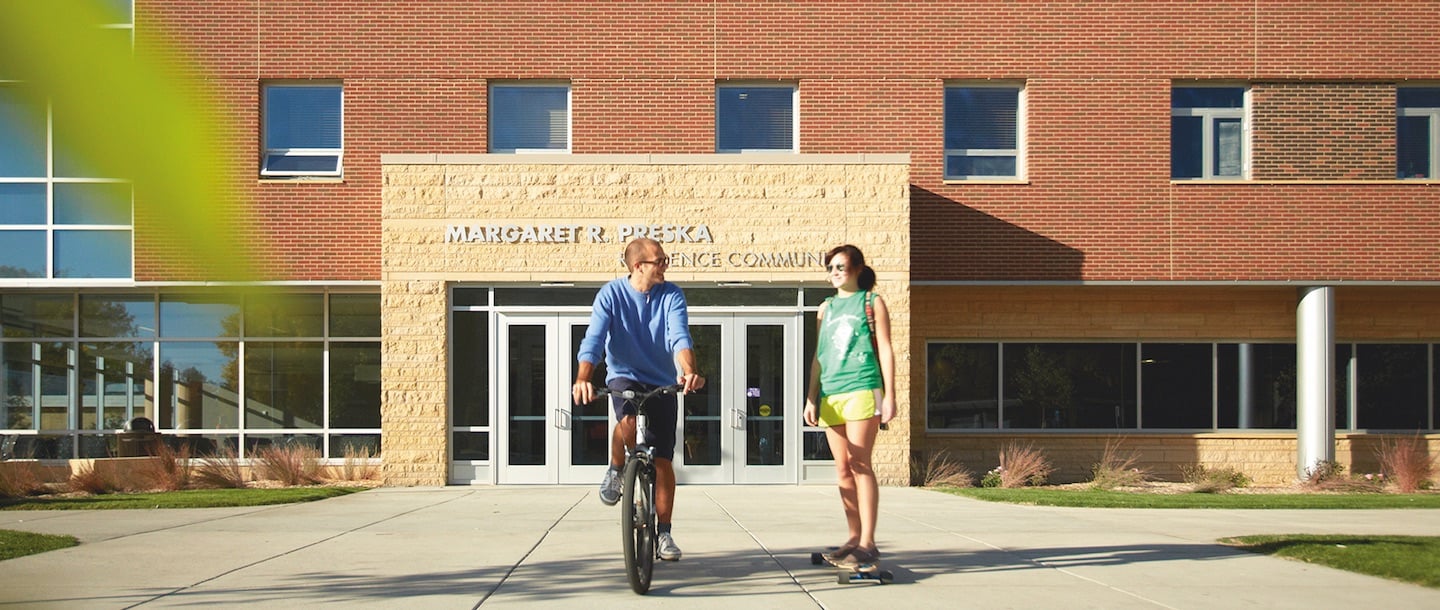 A boy bicycling and a girl skating in front of the Preska Community building