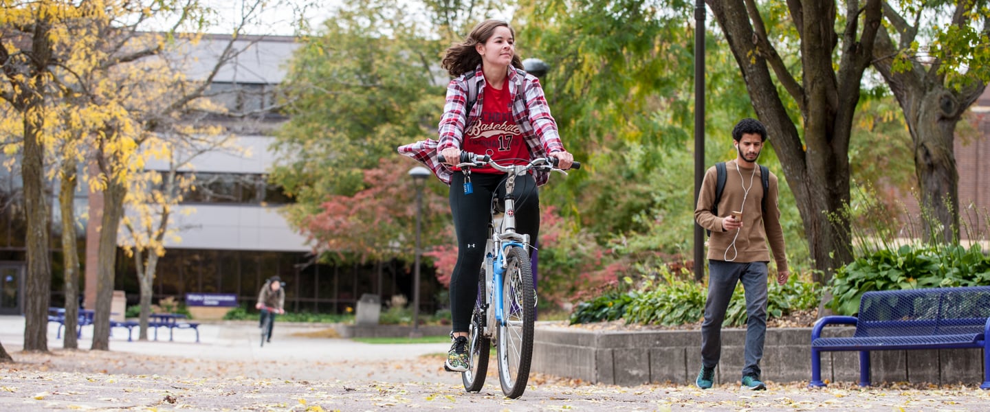 A student biking in front of the Wigley Administration building and another student walking listening to music with another student bicycling in the background on a fall day