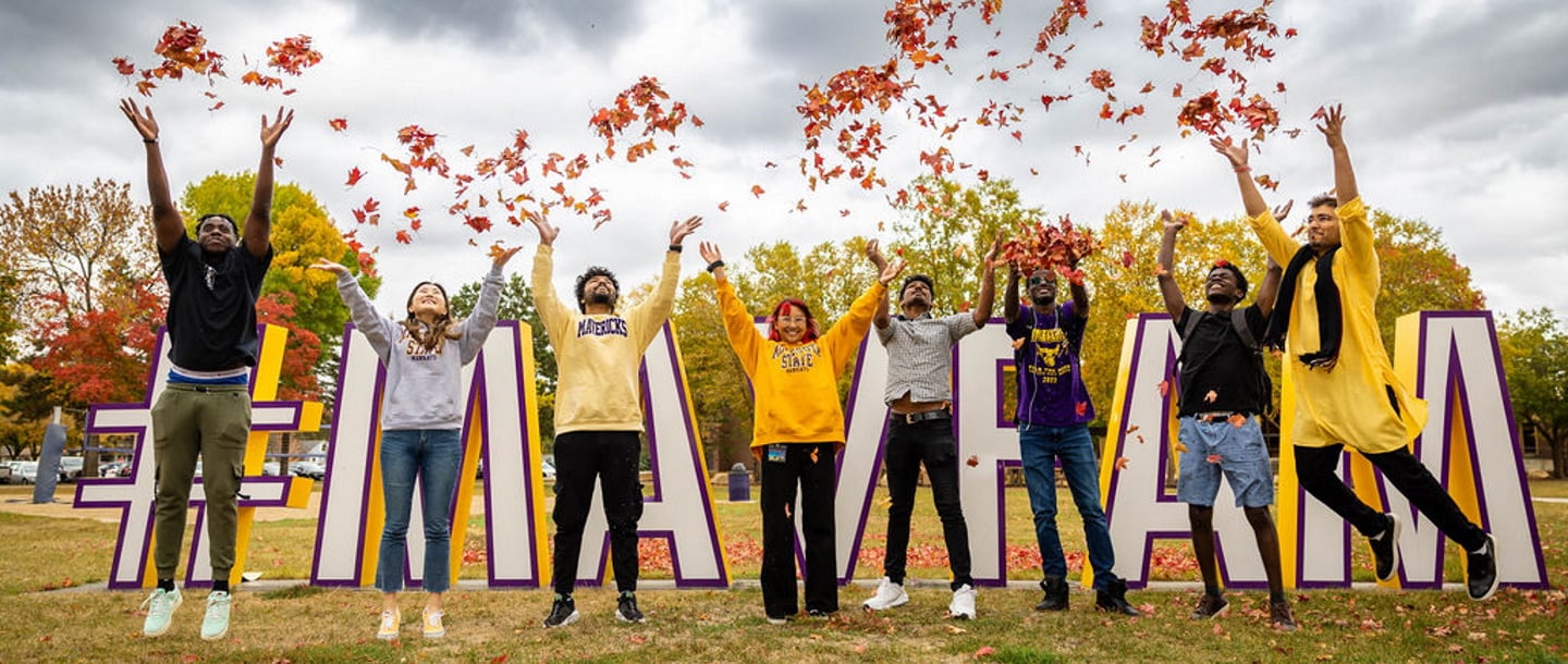 students throwing around the fallen leaves from trees with joy in front of the MAVFAM sign by the Performance Arts Center building