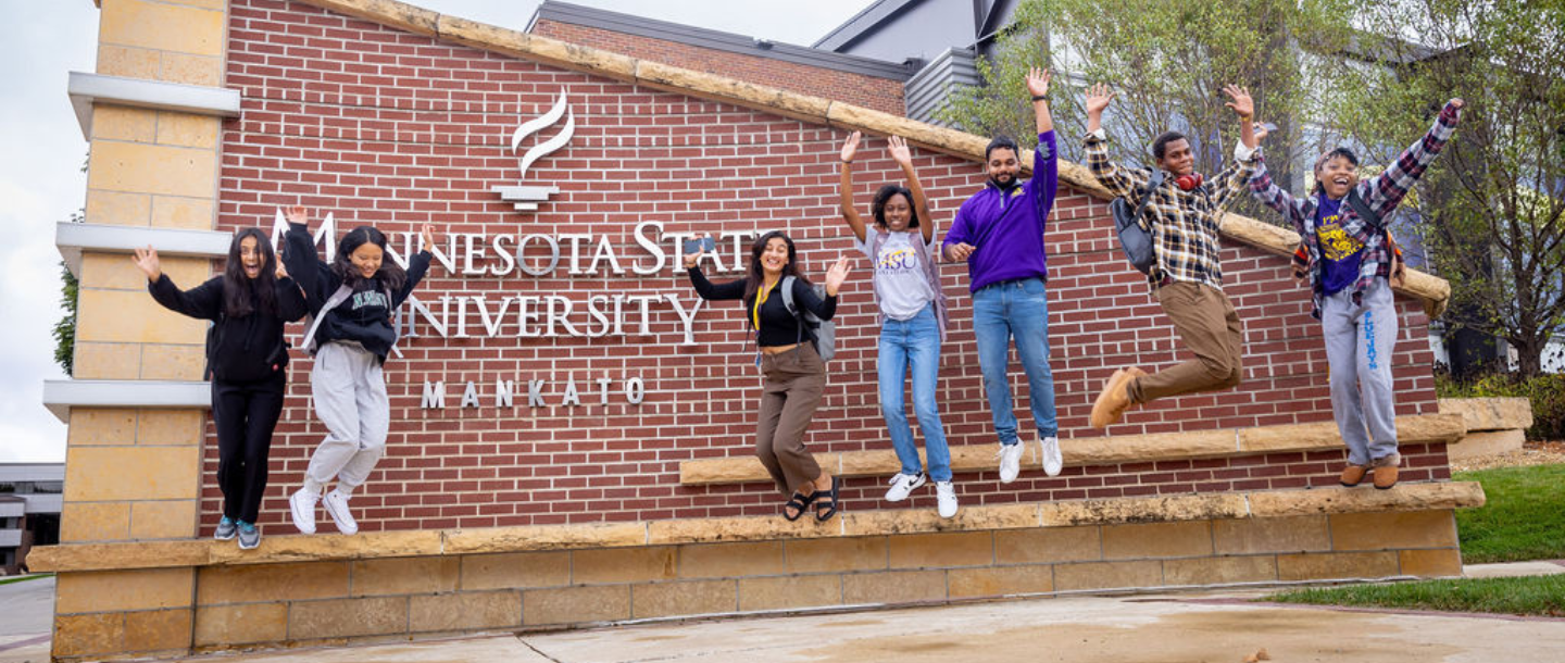 Seven students smiling as they are jumping off the bench by the Minnesota State University, Mankato entrance sign located in front of the Otto Recreation Center