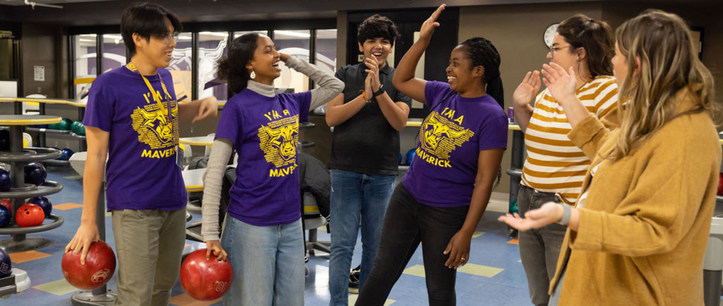 Six students in the Maverick Bullpen, two students are holding a bowling ball, smiling and clapping as two students are giving a high five