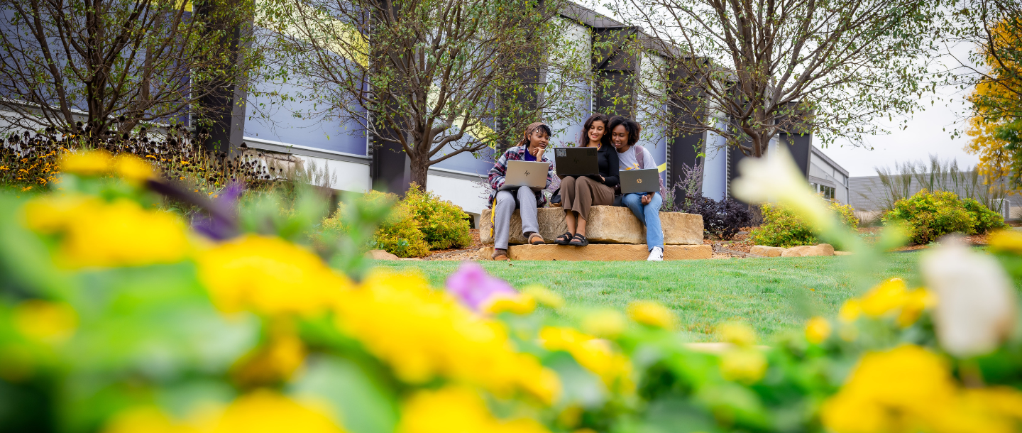 Three students sitting outside of campus on a stone bench with their laptops working together as a group