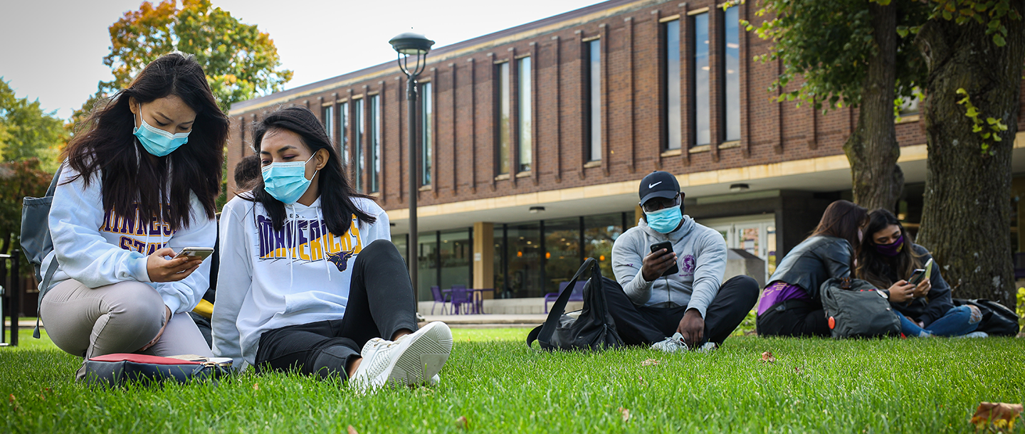 Students wearing masks sitting outside on the lawn behind the Centennial Student Union building