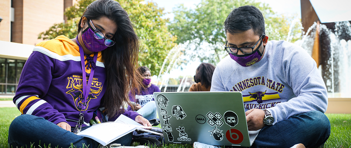 Students wearing masks sitting outdoors on the lawn by the fountain studying with a laptop
