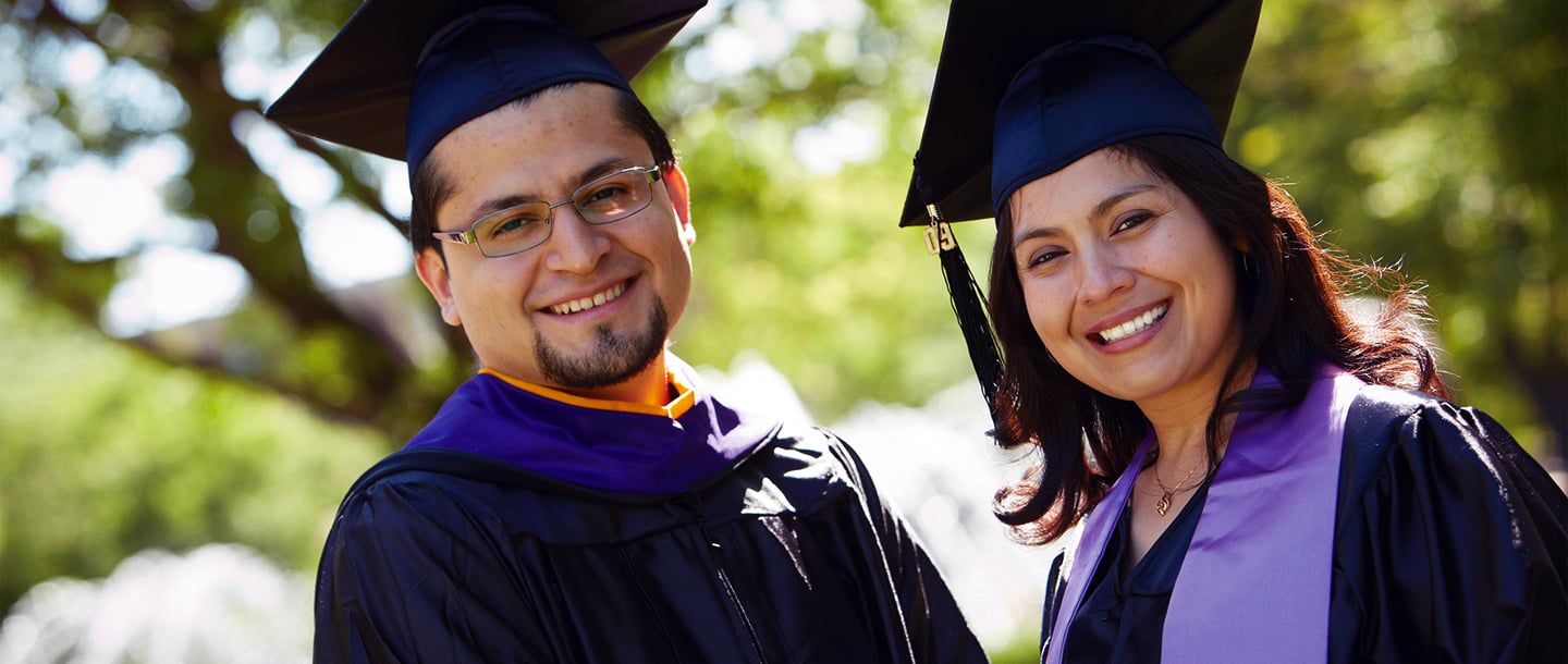 Two students posing outside of campus wearing black hats and gowns for graduation