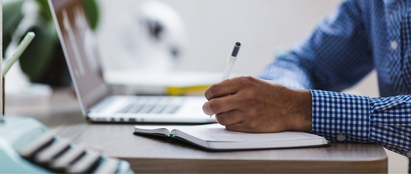 A person writing on a notebook at a desk with a laptop