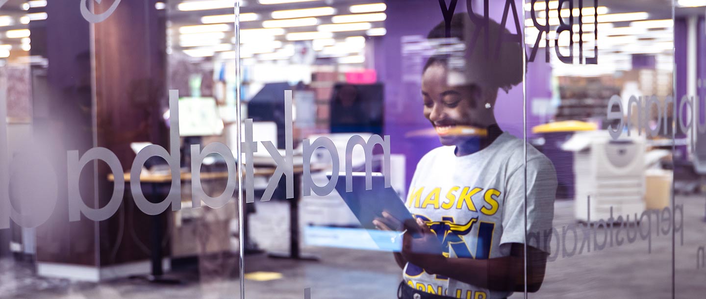 Photo of girl standing behind glass window in the Memorial Library. She is holding a tablet and smiling at it.
