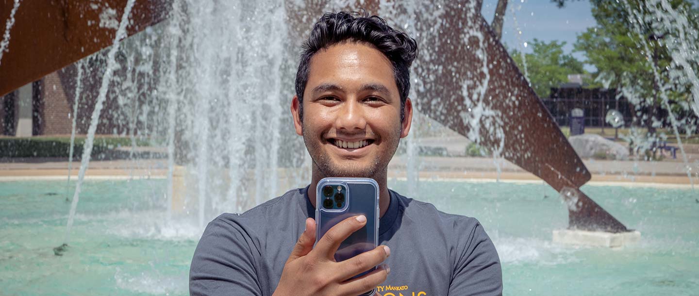 Guy sitting in front of a water fountain on campus holding his phone up and taking a selfie while smiling