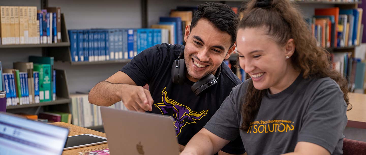 Two students sitting in the library working on a laptop and smiling and pointing at the screen