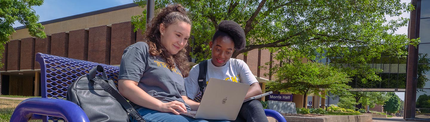 Two students sitting outside on a bench looking at a laptop together