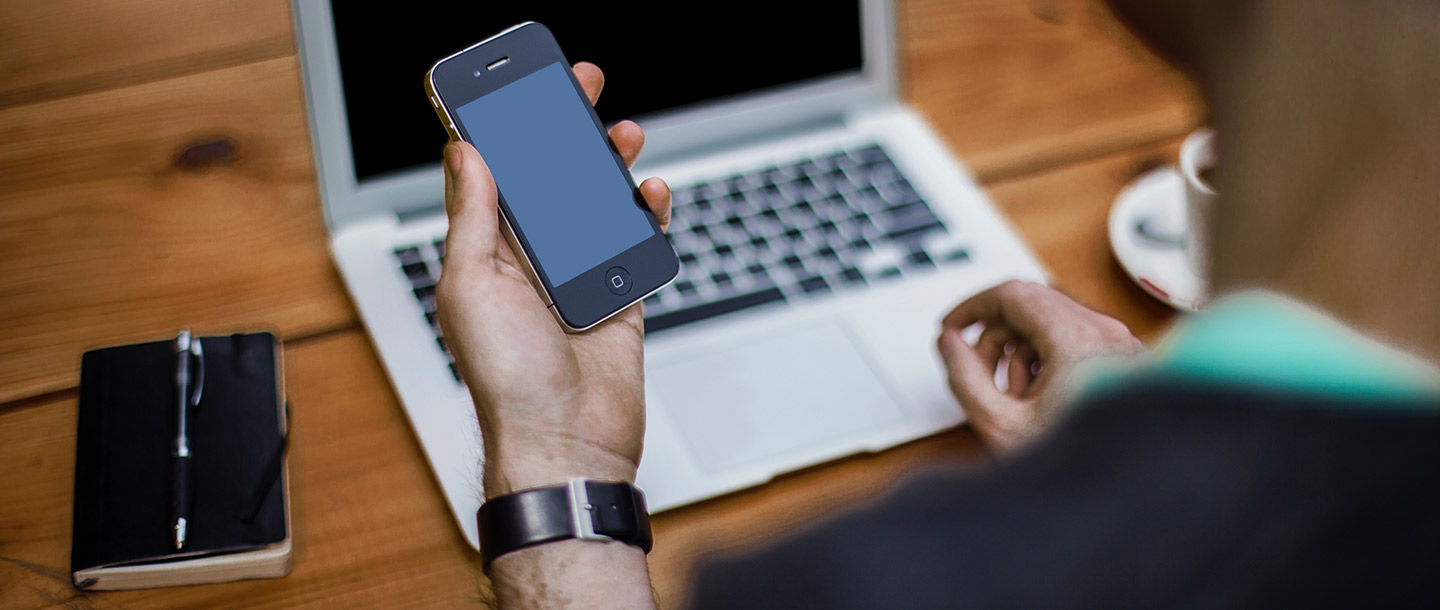 Staff member working remotely at a table with laptop and holding a smart phone