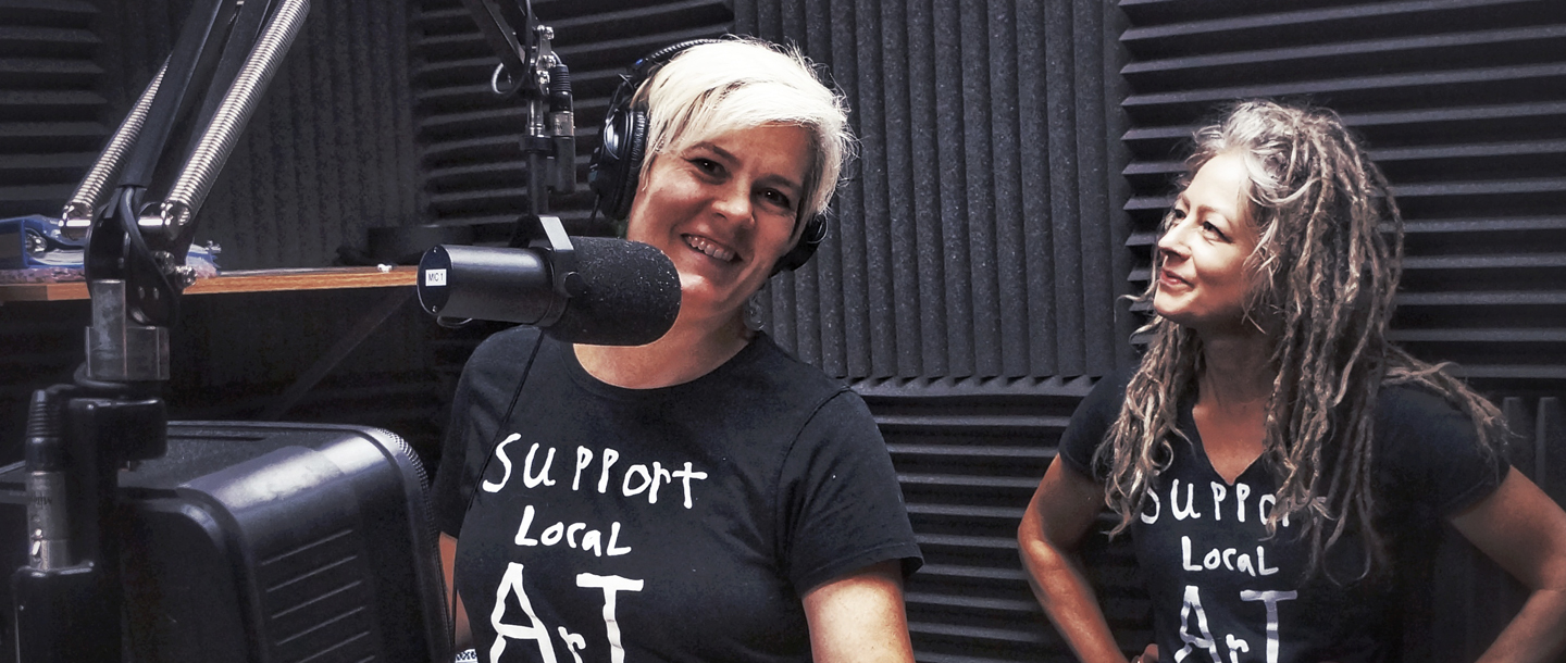 Two artists wearing black t-shirt saying "support local art", one in front of a speaker and another staring at the other artist at Header Art Center