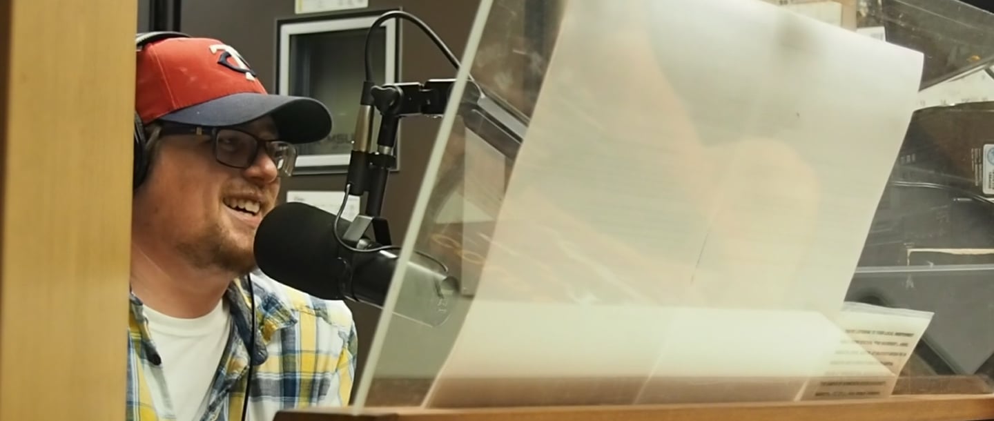 Eric "E Lars" Larson with a headphone on speaking into the microphone inside of the KMSU Radio station studio