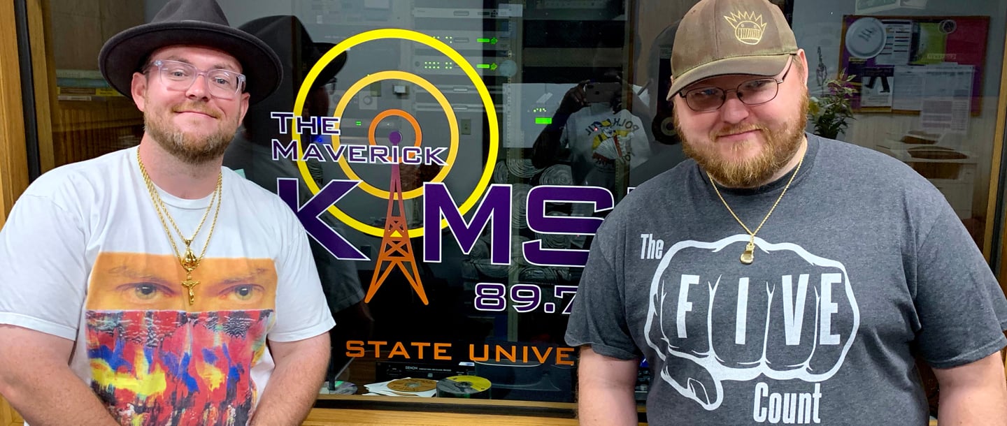 Juston "Ton" Cline and Dustin Wilmes posing and smiling infront of the studio window inside the KMSU Radio station