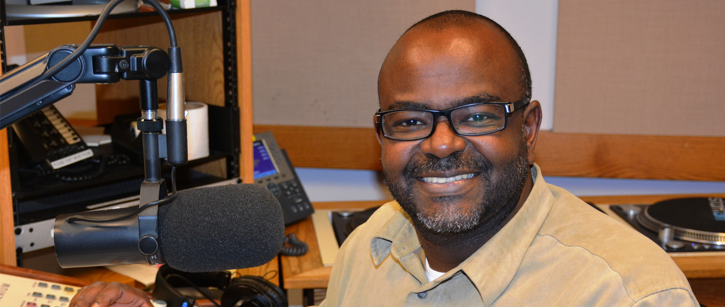 Mohamed Alsadig in front of microphone posing and smiling inside the KMSU Radio station studio