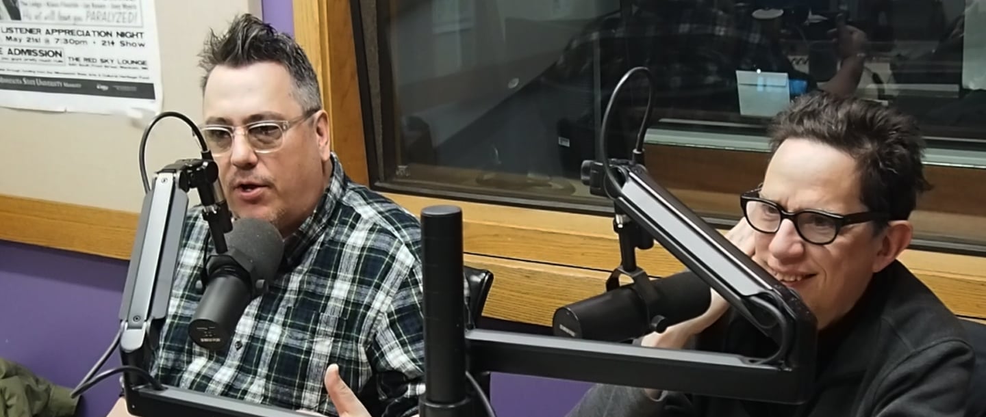 Two people one with a checkered shirt on and another person with a black seat shirt on speaking into microphones inside the KMSU Radio station studio