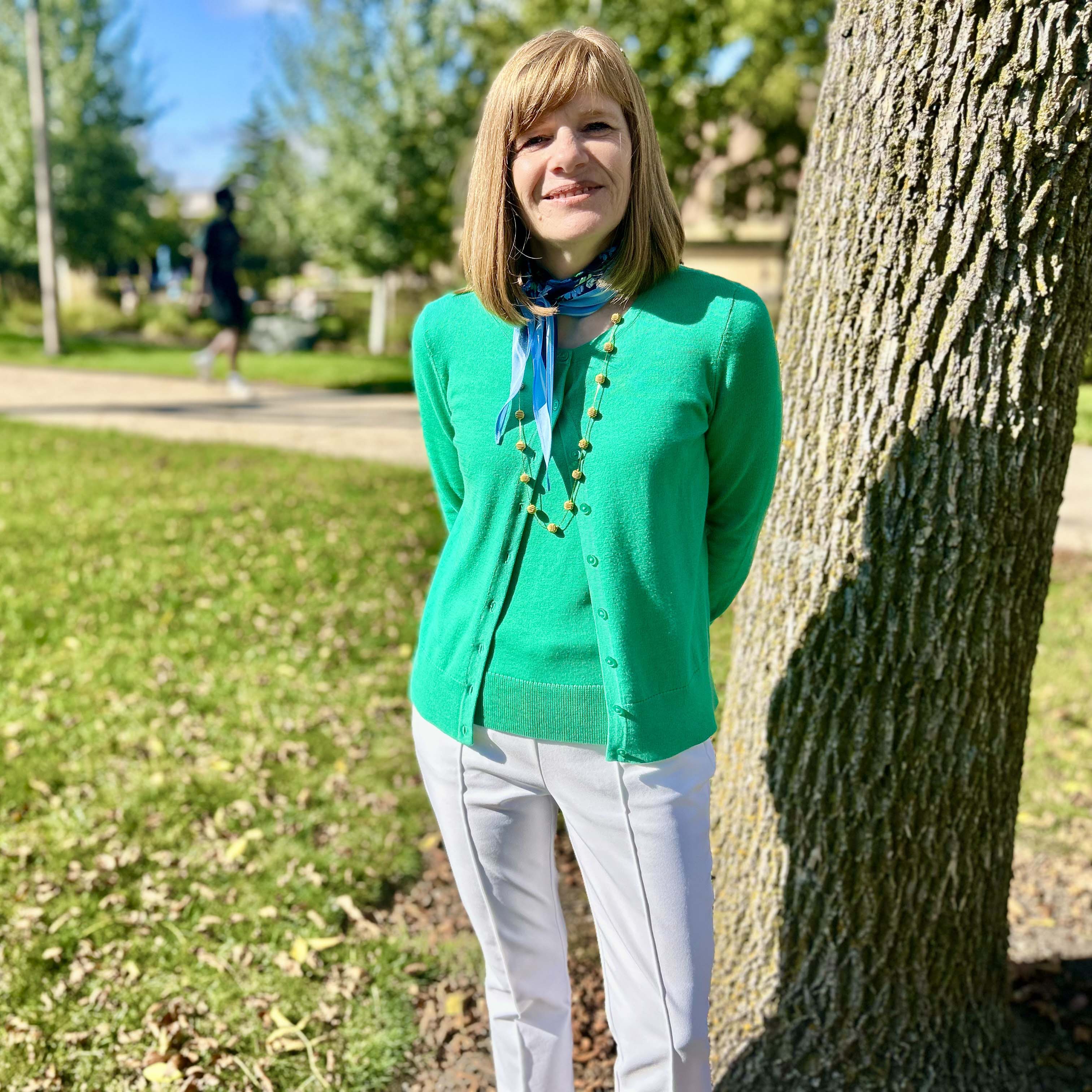 Nancy Fitzsimons standing outside near a tree in the fall sun. Nancy has on an emerald green top and white pants. Nancy is smiling at the camera with her hands behind her back.