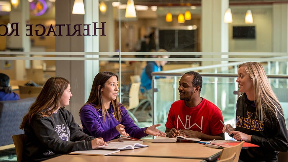Four students sitting together at a table in a common area on campus talking and working. 
