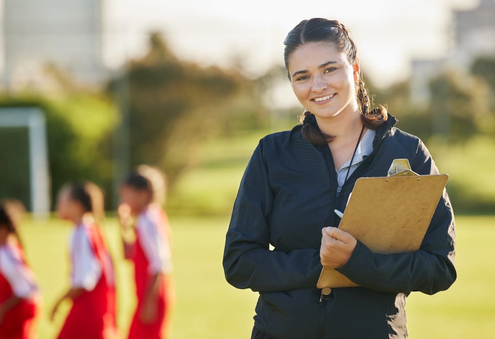 School gym teacher holding clip board, on the field with students.