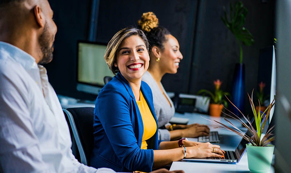 woman on laptop smiling at the male on laptop next to her. Both in business attire