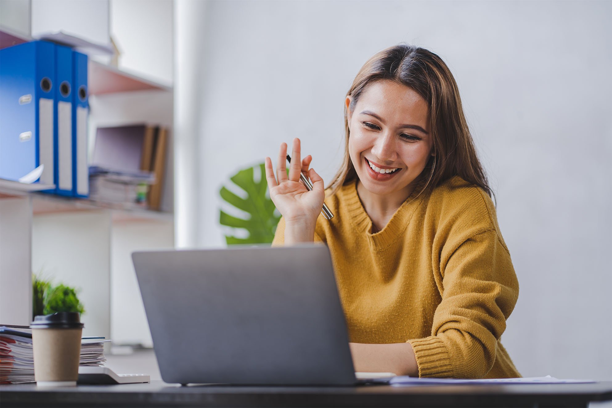 Student sitting at a desk, looking at her computer with her hand up and smiling.