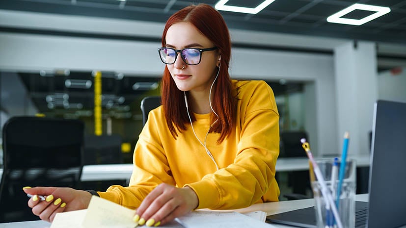 female writing in a notebook with laptop computer and pencils nearby