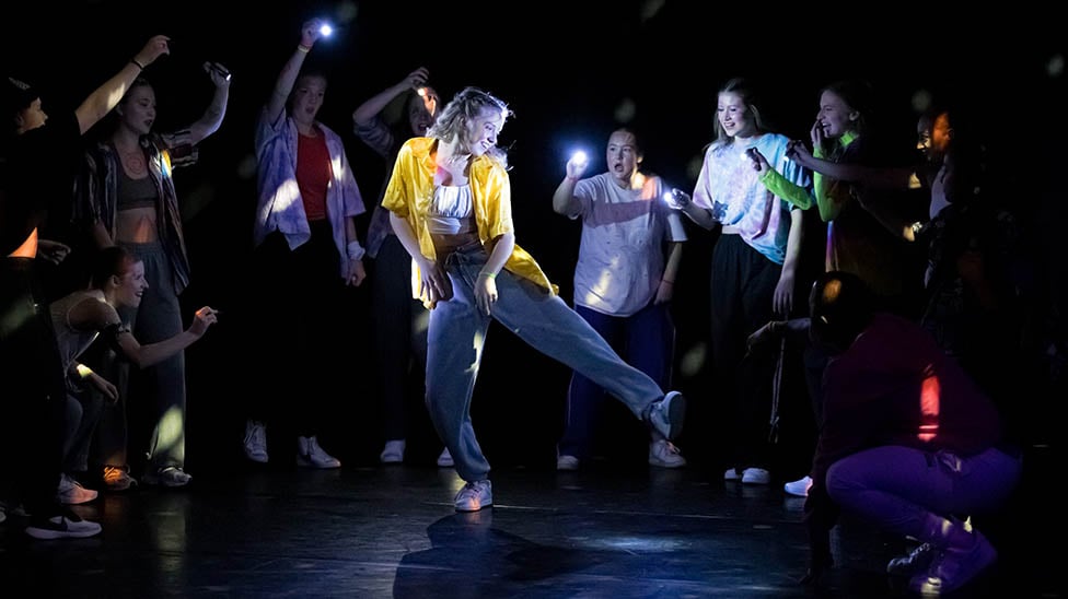 Students dancing on a dark stage with the spotlight on one dancer dressed in jeans and a tee