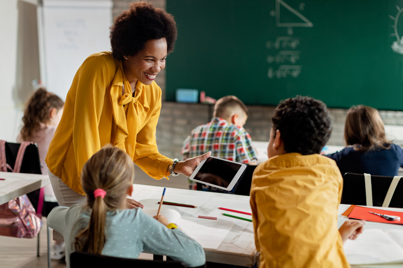 Teacher holding digital device helping students in classroom.