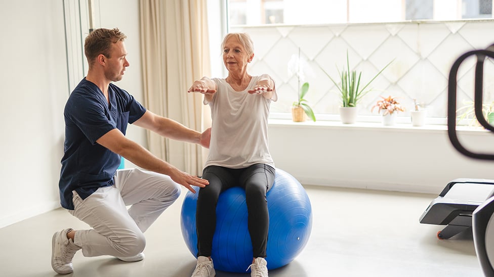 man helping older woman balance on a balance ball for therapy