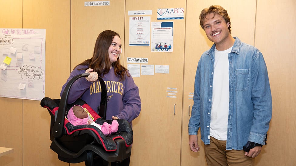 Two students in a FCS classroom working with a carseat and a pretend baby and bottle.
