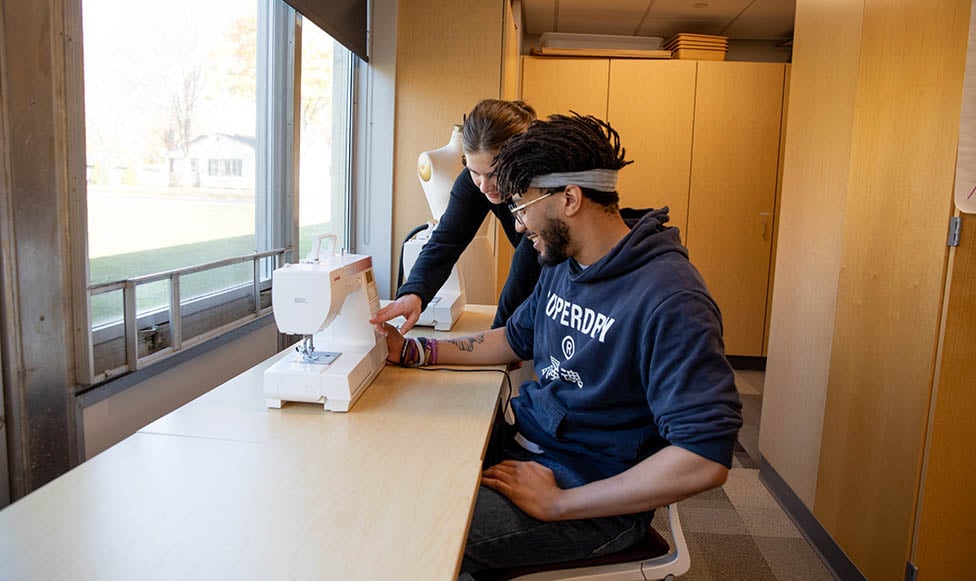 Male student sitting at the sewing machine in the textile lab while another student is helping with running the machine. 
