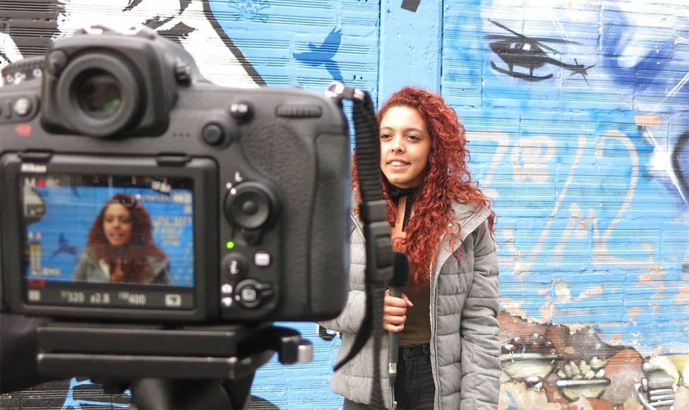 Film and Media student practicing in front of the camera. A beautiful painted mural is behind her and she is holding a microphone talking into the camera.