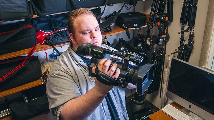 Male student holding up a camera looking through the finder with other tech equipment around.