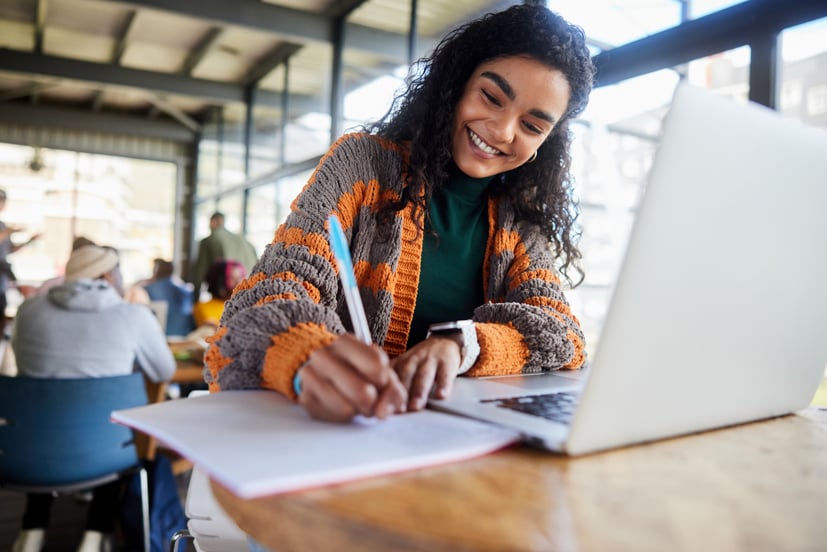 Woman sitting on at a table with computer in front of her and writing in a notebook.