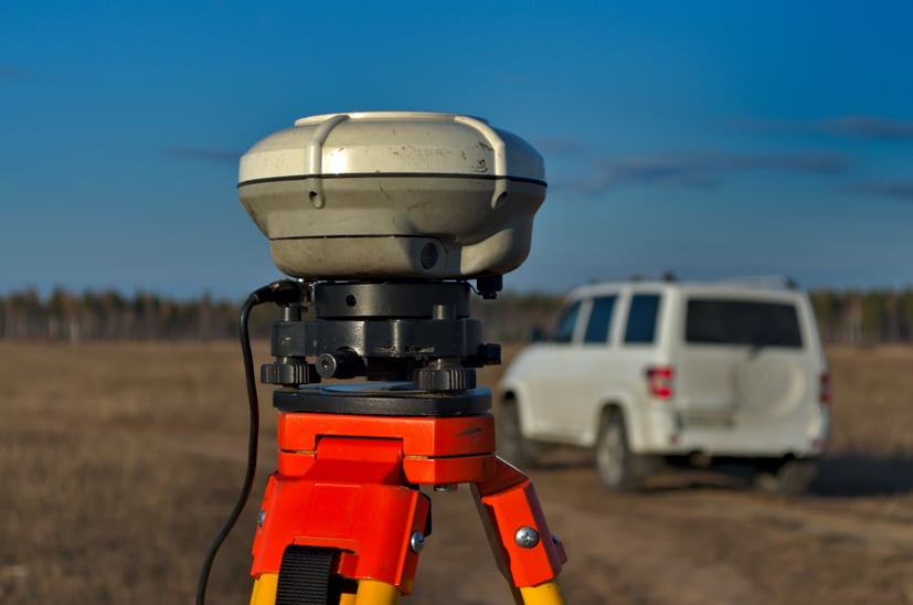 Survey tool on tripod in an open field with white vehicle in background. 