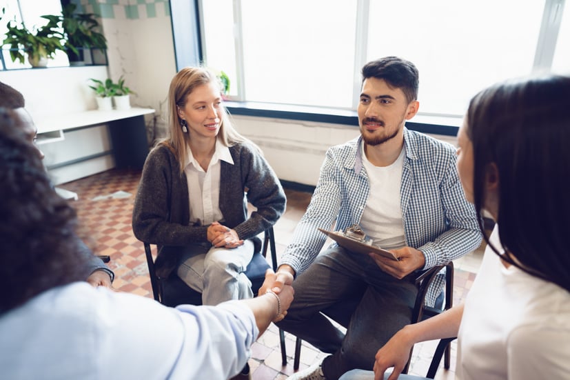 Man shaking hands with person, holding a clipboard, sitting in a circle. 