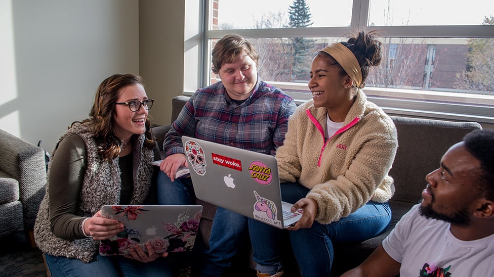 Students hanging around with a couple of laptops. Some sitting on the couch, one in a chair, and one on the floor.